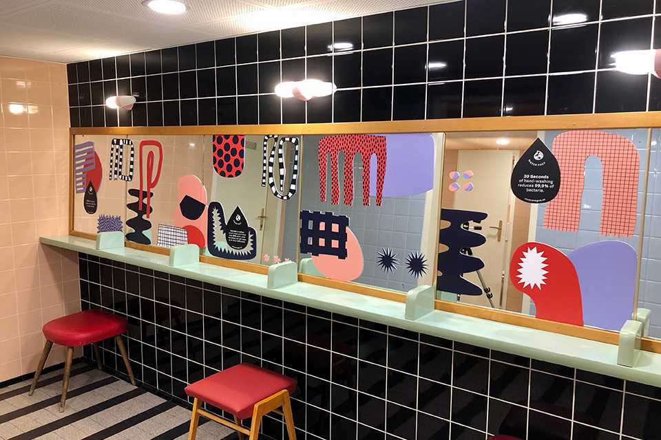 VIVA CON AGUA Toilette bei FORWARD: Buntes Design mit Spiegel und Hockern. Schwarze und rosa Fliesen.